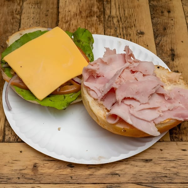 An open-faced bagel sandwich with ham, cheese, lettuce, and onions on a white paper plate.
