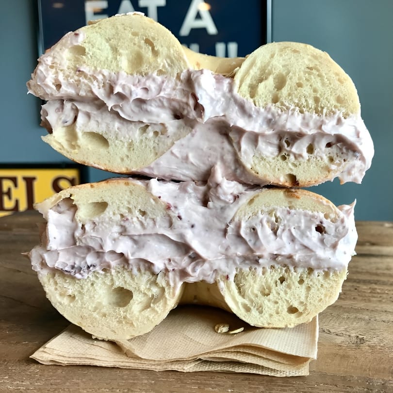 A Bagel Culture bagel, sliced in half, generously filled with purple cream cheese on a wooden table.