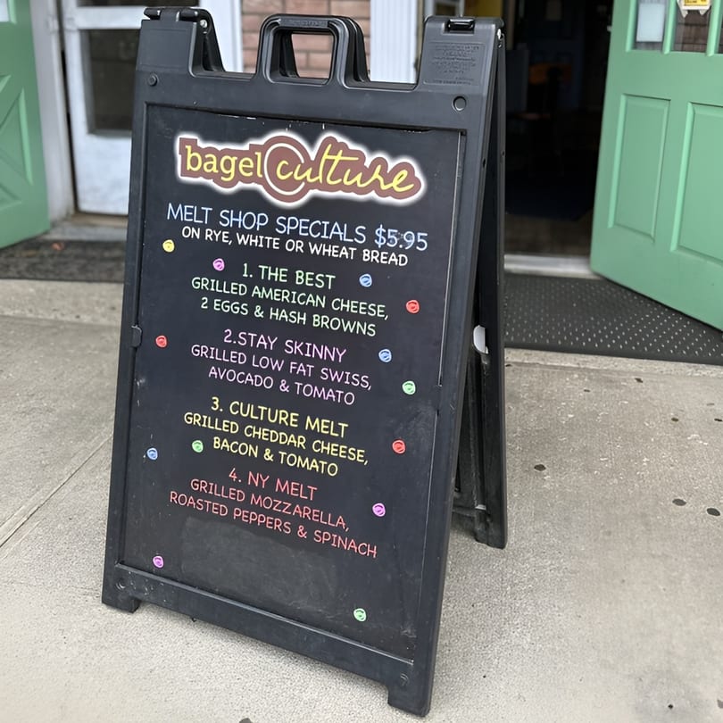 A black A-frame sign for Bagel Culture displays "Melt Shop Specials" with various grilled cheese options.