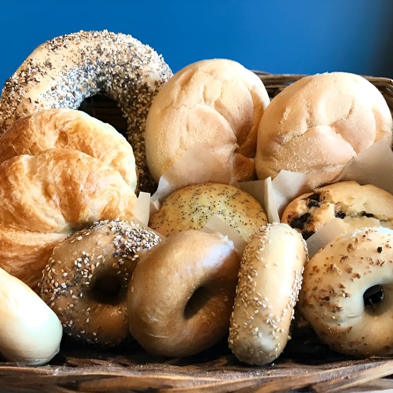 A rustic basket filled with various Bagel Culture bagels, rolls, and muffins against a blue background.