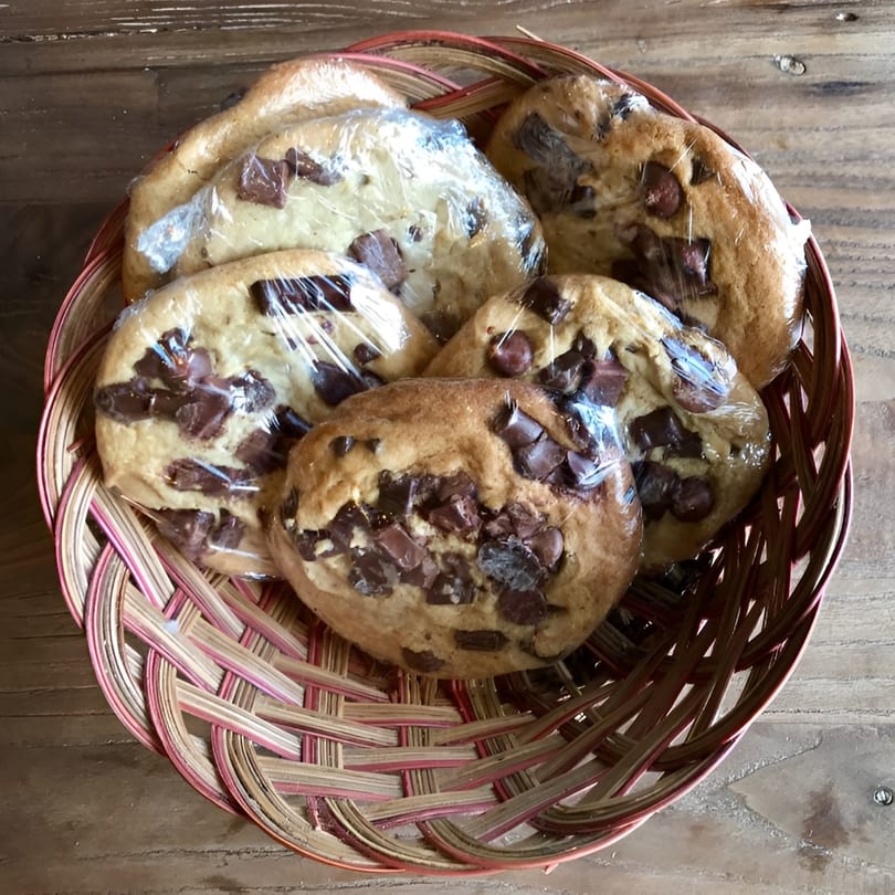 Several chocolate chip cookies, individually wrapped in plastic, fill a woven basket on a wooden surface.